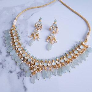 May include: A gold necklace and earrings set with light blue and clear stones, and white pearls. The necklace has a delicate chain with a pendant of light blue stones and white pearls. The earrings are drop earrings with a similar design.