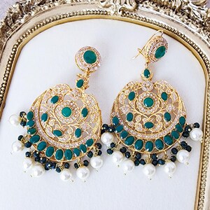 May include: A pair of gold-toned earrings with a circular design. The earrings feature emerald green gemstones and small clear crystals. White and black beads dangle from the bottom of the earrings. The earrings are displayed on a gold-framed surface.