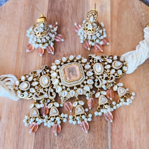 May include: A gold and white choker necklace with a large, square, peach-colored stone in the center. The necklace is decorated with smaller white stones and has peach-colored beads hanging from it. The necklace is paired with matching earrings.