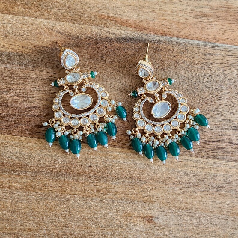 May include: Gold-toned chandelier earrings with clear and emerald green accents. The earrings feature a teardrop-shaped top, a central oval stone, and dangling green beads. Small white beads and clear stones add sparkle.