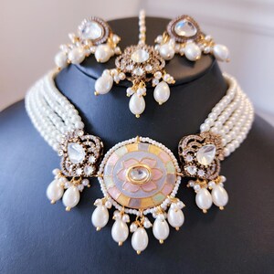 May include: A multi-strand pearl choker necklace with a large, round pendant featuring a floral design in pastel colors and rhinestones. The pendant is surrounded by smaller pearl drops. The set also includes a matching pair of earrings and a tikka.