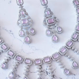May include: A silver necklace and earrings set with pink stones. The necklace has a delicate chain with a pendant featuring a row of rectangular stones and dangling teardrop stones. The earrings have a similar design with a smaller pendant and teardrop stones.