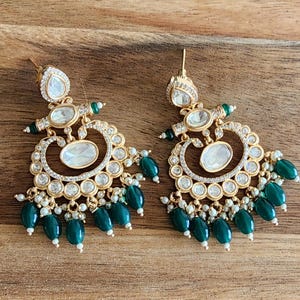 May include: Gold-toned earrings with clear and emerald green accents. The earrings feature a teardrop-shaped top, a central oval design, and a series of green bead drops. The earrings are set against a wooden surface.