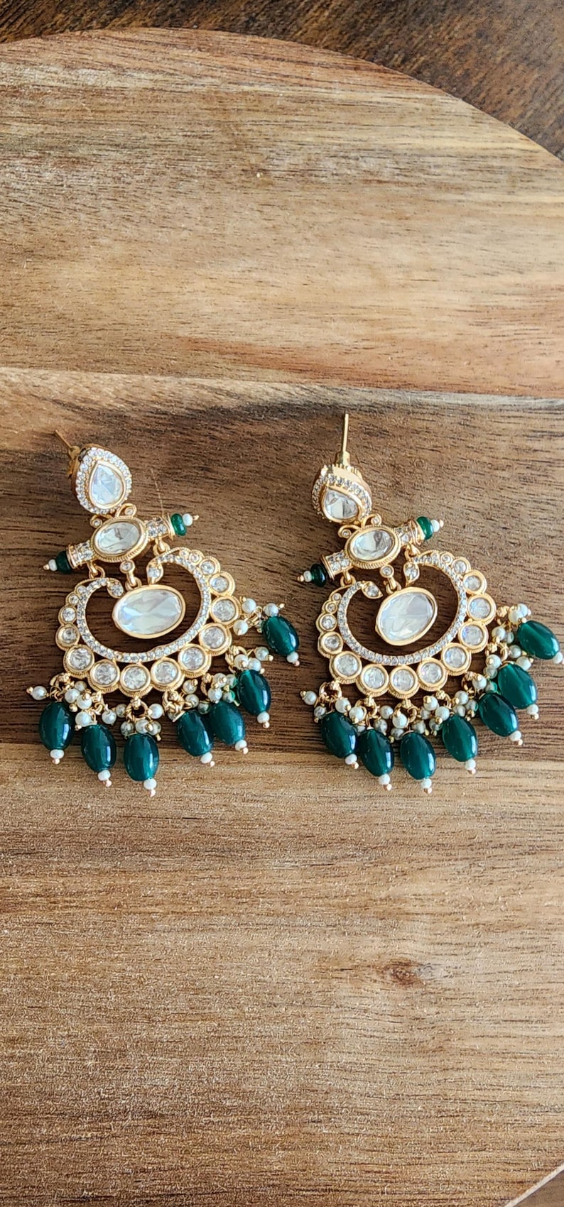 May include: Gold-toned earrings with clear and emerald green accents. The earrings feature a teardrop-shaped top, a central oval design, and a series of green bead drops. The earrings are set against a wooden surface.