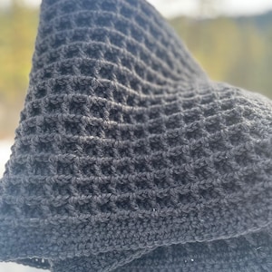 Puede incluir: Una bufanda de punto de waffle negro con un patrón texturizado. La bufanda está doblada por la mitad y drapeada sobre un poste de madera.