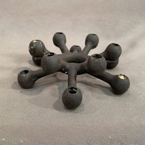 May include: A dark gray, star-shaped candle holder with seven circular openings. The candle holder has a matte finish and is made of a solid material. The design is symmetrical, with each opening positioned around a central ring.