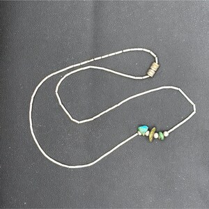 May include: A delicate necklace featuring a thin, silver-toned chain. The necklace is adorned with small, turquoise-colored stones and other green and white beads. The clasp is a small, silver-toned magnetic closure. The necklace is displayed on a dark surface.
