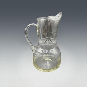 May include: A clear glass pitcher with a handle and a spout. The pitcher has a round base and a cylindrical top. Etched starburst designs decorate the top portion. The pitcher is likely used for serving beverages.