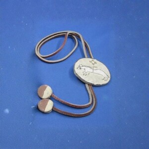 May include: A bolo tie with a silver-colored oval pendant featuring an etched design of an animal and a sun symbol. The tie has a brown leather cord and two round, two-tone brown and white tips. The background is a solid blue.