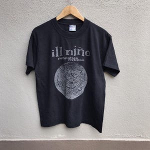 以下が含まれることがあります： バンド名「ill niño」と「revolution revolución」の文字がプリントされた黒いTシャツ。円形のデザインの上に配置されています。デザインは詳細で様式化された画像が特徴です。Tシャツは木製のハンガーに掛けられています。