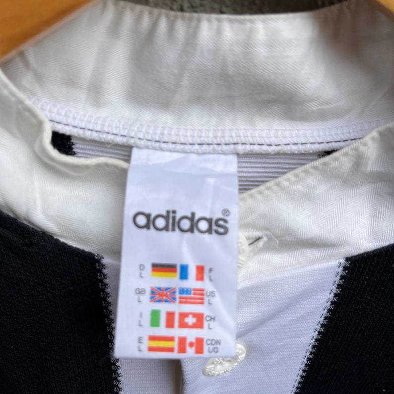 May include: Close-up of a white and black Adidas shirt collar. The Adidas logo is visible on a tag with country flags. The shirt has a white collar and a black and white striped design.