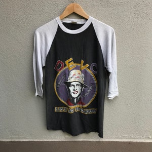 Op de afbeelding: Zwart-wit baseball-stijl t-shirt met een zwart-wit afbeelding van een man met een hoed en de tekst "DEVO ARE DEVO!" en "LIVE IN CONCERT".