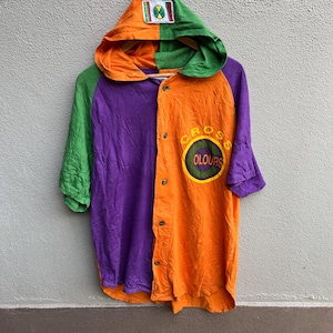 Puede incluir: Una colorida camisa con capucha y mangas cortas. La camisa está dividida verticalmente en paneles naranjas y morados, con mangas y capucha verdes. La capucha tiene un parche con un diseño verde y amarillo. La camisa presenta el texto "CROSS OLOURS" en un diseño circular.