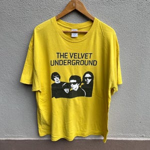 Könnte beinhalten: Senfgelbes T-Shirt mit dem Text "THE VELVET UNDERGROUND" und einem Schwarz-Weiß-Bild der Band. Das T-Shirt hängt an einem Holzbügel vor einem neutralen Hintergrund.