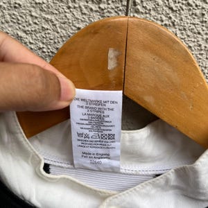 May include: A close-up of a white collared shirt with a black stripe detail. The shirt tag displays text in multiple languages, including "THE BRAND WITH THE 3 STRIPES" and "Made in England".