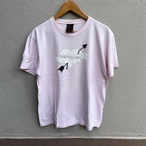 Könnte beinhalten: Hellrosa T-Shirt mit Rundhalsausschnitt und kurzen Ärmeln. Das Shirt zeigt eine Grafik eines von einem Pfeil durchbohrten Herzens mit den Worten "BLOOD RED SHOES" auf einem Banner. Das T-Shirt hängt an einem Holzkleiderbügel.
