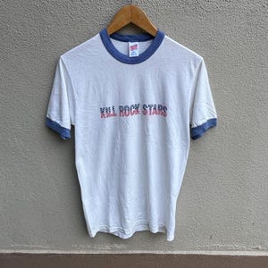 May include: White t-shirt with blue trim on the collar and sleeves. The text "KILL ROCK STARS" is printed across the chest in a red and blue design. The shirt is hanging on a wooden hanger.