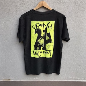 Puede incluir: Camiseta negra con un gráfico verde neón en la espalda. El gráfico presenta las palabras "GRIND 24 VIOLENT" en una fuente desgastada, junto con imágenes de naipes. La camiseta está colgada en una percha de madera.