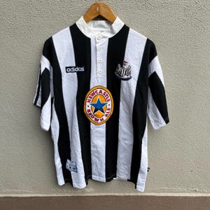 May include: Black and white striped soccer jersey with a white collar and button-up placket. Features the Adidas logo, a team crest, and a Newcastle Brown Ale logo. The jersey is short-sleeved.