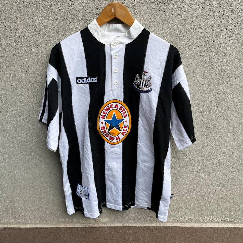 May include: Black and white striped soccer jersey with a white collar and button-up placket. Features the Adidas logo, a team crest, and a Newcastle Brown Ale logo. The jersey is short-sleeved.