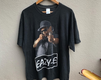 May include: Black t-shirt with a graphic of a person in a hat and sunglasses, aiming two guns. The text "EAZY-E" is in a white box. The shirt is displayed on a wooden hanger against a neutral background.