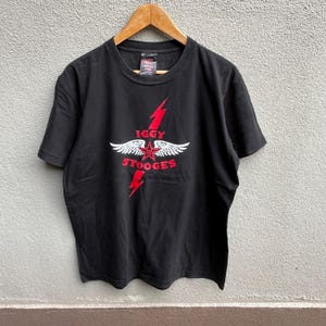 Könnte beinhalten: Schwarzes T-Shirt mit roter und weißer Grafik mit dem Text "IGGY STOOGES", einem roten Blitz, einem Stern und Flügeln. Das T-Shirt hängt an einem Holzkleiderbügel.