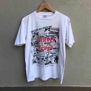 May include: White t-shirt featuring a collage of newspaper clippings with the words "Jungle Fever" in red. The shirt also includes the text "A Spike Lee Joint" at the bottom. The design is a mix of black and white with red accents.