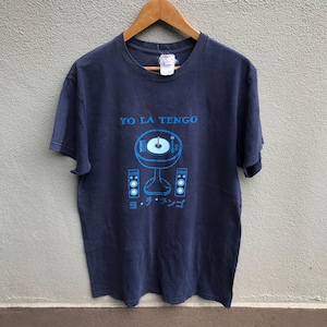 Puede incluir: Camiseta azul marino con un gráfico azul de un tocadiscos y altavoces. El texto "YO LA TENGO" está encima del gráfico, con texto japonés debajo. La camiseta está colgada en una percha de madera.