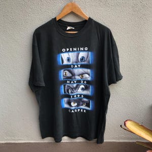 Pode incluir: Camiseta preta com um gráfico de Casper, o Fantasma Amigável. O texto diz "OPENING DAY MAY 26 1995 CASPER". O gráfico mostra os olhos de Casper em uma moldura azul.