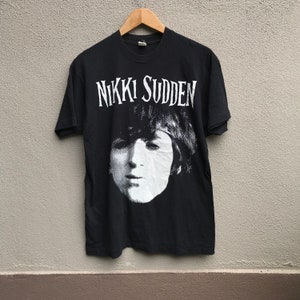May include: Black t-shirt with a white graphic of a person's face and the text "Nikki Sudden" printed on the front.