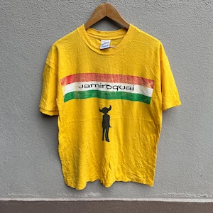 Könnte beinhalten: Gelbes Jamiroquai-T-Shirt mit horizontalem Streifendesign in Rot, Weiß und Grün. Der Name der Band ist weiß gedruckt. Eine schwarze Silhouette einer Figur mit Hörnern befindet sich darunter.