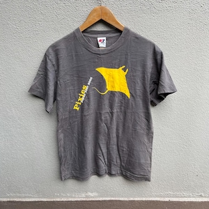 Puede incluir: Camiseta gris con un diseño de manta raya amarilla. El texto "Pixies Japan Tour 2005" está impreso en amarillo. La camiseta está colgada en una percha de madera. La tela está arrugada.