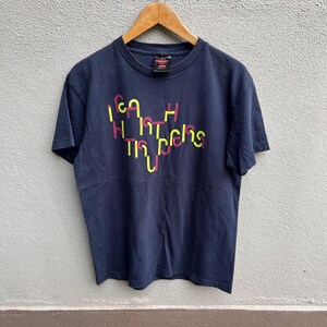 May include: Navy blue t-shirt with a graphic design in yellow and pink. The text on the shirt reads "Cash Hustlers" in a stylized font. The shirt is hanging on a wooden hanger.