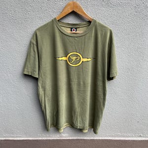 Puede incluir: Camiseta verde oliva con un gráfico amarillo y negro en la parte delantera. El gráfico presenta una letra "P" estilizada dentro de un círculo, con formas de rayo a cada lado. La camiseta está colgada en una percha de madera.