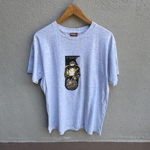 Puede incluir: Camiseta gris claro con un gráfico de una figura con máscara de soldadura y la inscripción "Santa Cruz". La camiseta está colgada en una percha de madera.