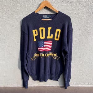 May include: Navy blue sweater with the word "POLO" in yellow block letters above an American flag graphic. Below the flag is a banner with "RALPH LAUREN" in yellow script. The sweater is hanging on a wooden hanger.