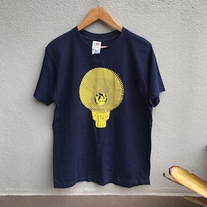 Puede incluir: Camiseta azul marino con un diseño amarillo de una bombilla con líneas radiantes y el texto "STEREO LAB". La camiseta está colgada en una percha de madera. Un diseño llamativo.