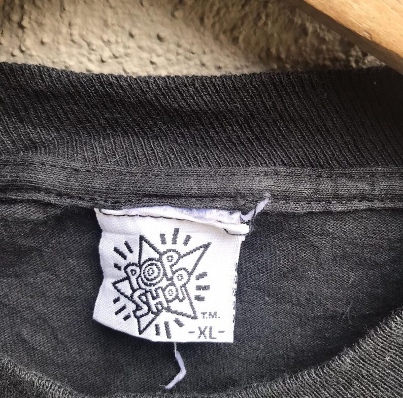 Puede incluir: Primer plano de un cuello de camiseta gris oscuro con una etiqueta blanca. La etiqueta presenta el texto "POP SHOP" en un dise&ntilde;o de estrella, con "T.M." y "-XL-" impresos debajo. La camiseta parece ser vintage.