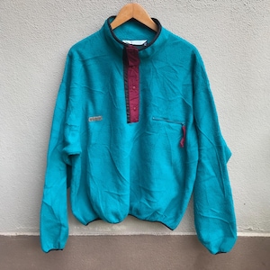 Op de afbeelding: Een turquoise blauwe fleece trui met een bordeauxrode drukknoopsluiting en een klein logo-patch op de linker borst. De fleece heeft lange mouwen en een ontspannen pasvorm.