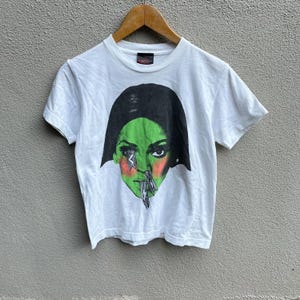 Peut inclure: T-shirt blanc avec un graphisme d'un visage de femme à la peau verte, aux cheveux noirs et à un motif d'éclair. Le t-shirt a un col rond et des manches courtes. Le nom de la marque "MOO" est imprimé sur le col.