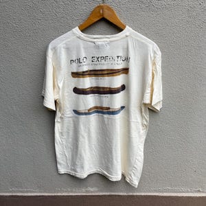 May include: Cream-colored t-shirt with the text "POLO EXPEDITION" and illustrations of three canoes. The shirt features the words "SURVEY EQUIPMENT & BOAT" above the canoes and the names of the canoes below them.
