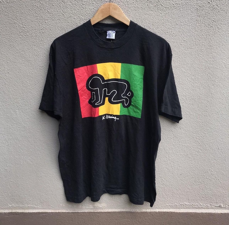 Puede incluir: Camiseta negra con un dise&ntilde;o gr&aacute;fico colorido con bloques rojos, amarillos y verdes, y un contorno blanco de una figura que gatea. La camiseta incluye el texto "K. Haring..." en blanco. La camiseta est&aacute; colgada en una percha de madera.