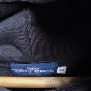 Pode incluir: Uma camisola com capuz preta com uma etiqueta interior azul no colarinho com o texto branco Tokyo Disney Resort e uma pequena etiqueta branca separada com a letra M indicando o tamanho, vis&iacute;vel no tecido escuro da pe&ccedil;a.