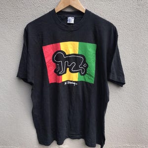 Puede incluir: Camiseta negra con un dise&ntilde;o de K. Haring. El dise&ntilde;o presenta un contorno blanco de una figura que gatea sobre un fondo rojo, amarillo y verde. La firma del artista est&aacute; debajo.