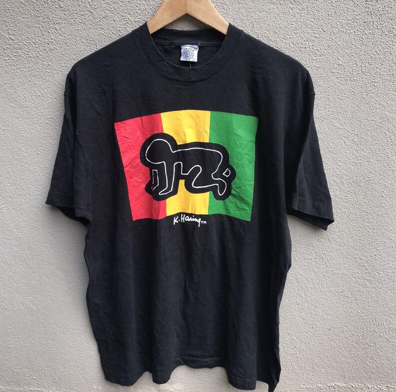 Puede incluir: Camiseta negra con un dise&ntilde;o de K. Haring. El dise&ntilde;o presenta un contorno blanco de una figura que gatea sobre un fondo rojo, amarillo y verde. La firma del artista est&aacute; debajo.