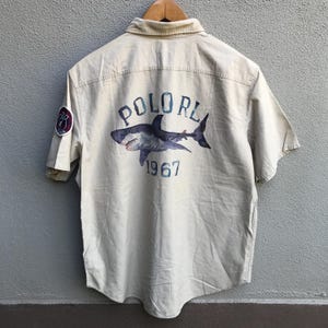 Puede incluir: Una camisa de manga corta beige con botones con un gráfico de tiburón blanco y el texto "POLO RL 1967" en la espalda.