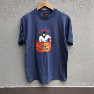 Pode incluir: Camiseta azul marinho com um desenho animado de Space Ghost em um barril. O texto "Alrighty" está acima do desenho e "Space Ghost Coast to Coast" abaixo. A camiseta está pendurada em um cabide de madeira.