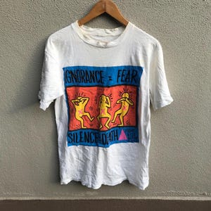 Pu&ograve; includere: Maglietta bianca con una stampa grafica di tre figure stilizzate con le mani sulle orecchie. La grafica &egrave; in uno schema di colori blu, rosso e giallo e dice "Ignorance = Fear" e "Silence = Death". La grafica include anche il testo "Fight AIDS Act Up".