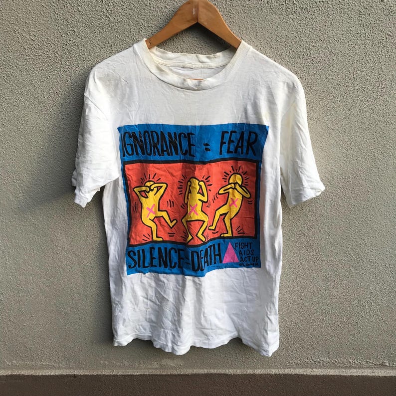 Pu&ograve; includere: Maglietta bianca con una stampa grafica di tre figure stilizzate con le mani sulle orecchie. La grafica &egrave; in uno schema di colori blu, rosso e giallo e dice "Ignorance = Fear" e "Silence = Death". La grafica include anche il testo "Fight AIDS Act Up".