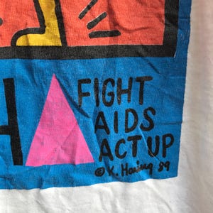 Pu&ograve; includere: Una maglietta bianca con una grafica blu e rosa. La grafica presenta un triangolo rosa, una figura nera e il testo "FIGHT AIDS ACT UP" con una firma che recita "&copy; K. Haring 89".